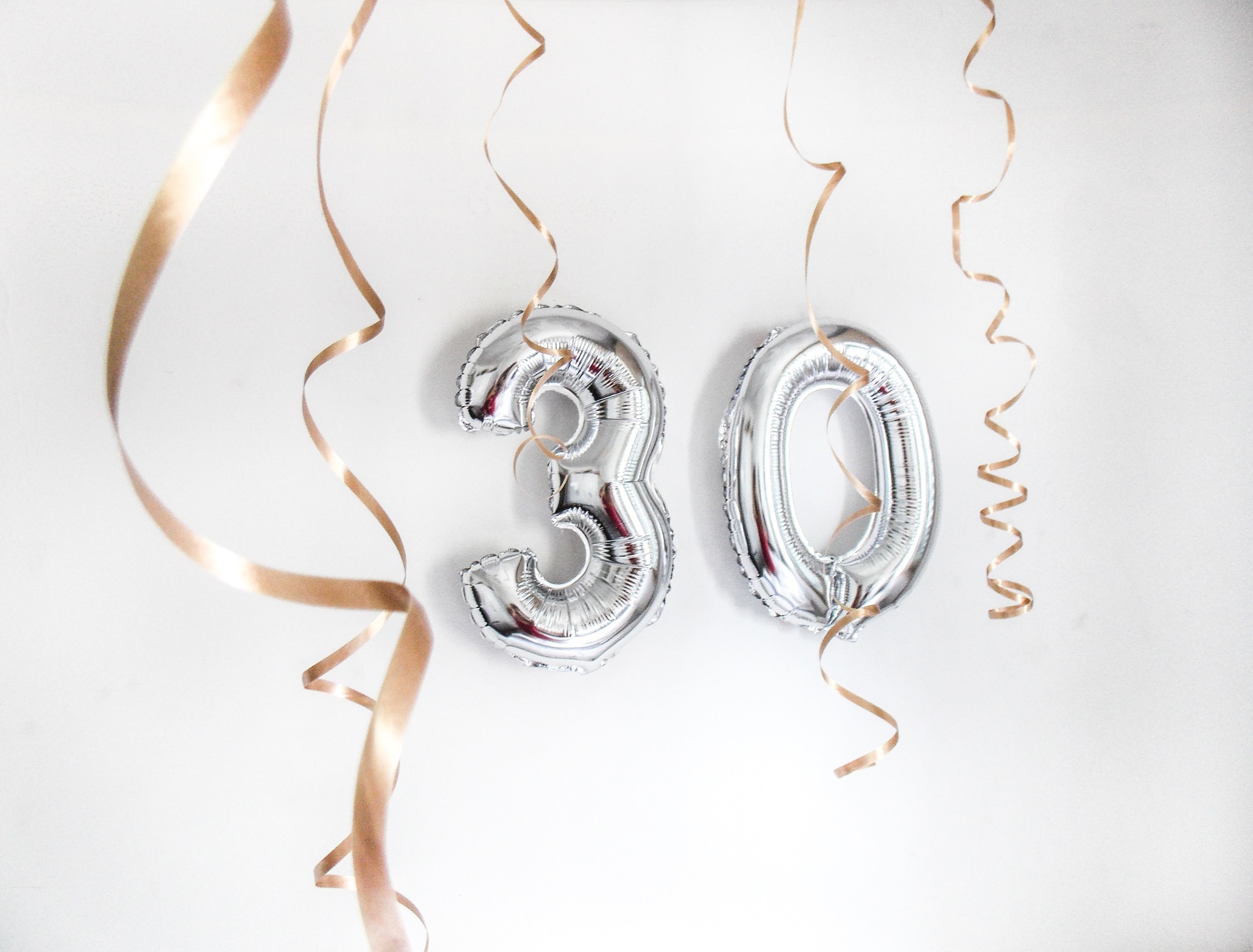 30th birthday celebration image of two silver balloons shaped as a three and zero, with gold spirals of ribbon floating above and through it. The image is from a free stock site called Unsplash and was taken by photographer Marina Barcelos.